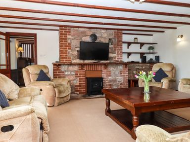 A living room with beige armchairs a wooden coffee table with flowers and a stone fireplace with a mounted TV at Valley House in Halberton near Tiverton
