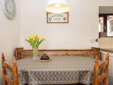 A dining area with wooden chairs and bench a table with grey patterned cloth yellow tulips in a vase and a covered dish at Valley House in Halberton near Tiverton