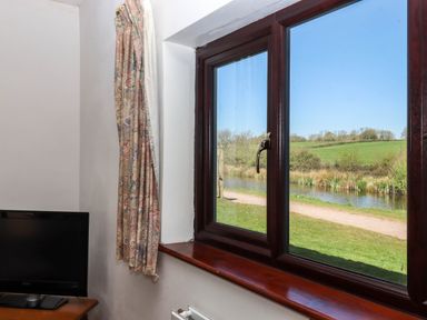 A window with floral curtains overlooking a river and green fields with a television on a wooden table at Valley House Halberton near Tiverton