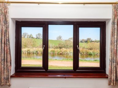 A window with dark wooden frame and patterned curtains overlooking a pond and green fields at Valley House in Halberton near Tiverton
