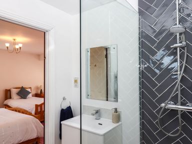 A bathroom with a sink and a shower area with gray tiles next to a bedroom with two wooden beds at Valley House in Halberton near Tiverton