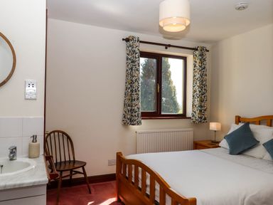 A bedroom with a wooden bed and bedside table a window with patterned curtains and a wooden chair at Valley House in Halberton near Tiverton