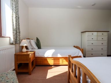 A bedroom with two wooden beds a wooden nightstand with a lamp a white chest of drawers and a window with patterned curtains at Valley House Halberton near Tiverton