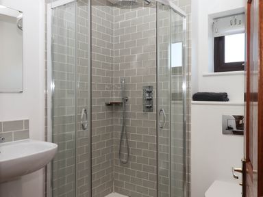 A bathroom with a glass enclosed shower with gray tiled walls a white sink and a small window with a black towel on the sill at Valley House in Halberton near Tiverton