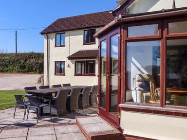 A patio with a long table and multiple chairs outside a two story house with large windows at Valley House in Halberton near Tiverton