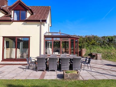 An outdoor patio with a dining table and chairs in front of a house with a conservatory at Valley House in Halberton near Tiverton