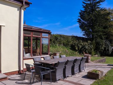 An outdoor patio with a long wooden table and wicker chairs next to a house with large windows at Valley House Halberton near Tiverton