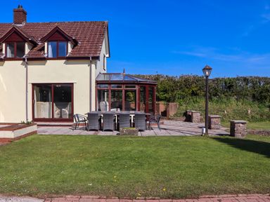 An outdoor patio with a dining table and chairs next to a house with a conservatory and a lawn at Valley House in Halberton near Tiverton