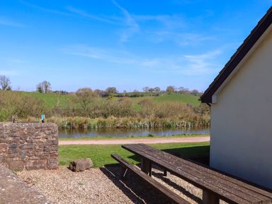 A picnic table next to a path with a stone wall and a river in a rural landscape at Valley House in Halberton near Tiverton