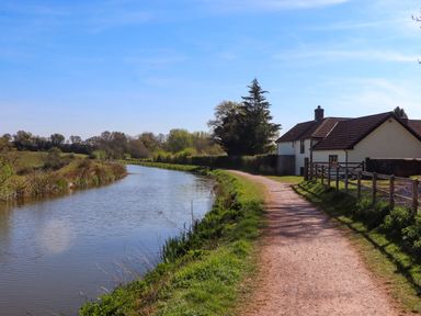 A dirt path beside a canal and a house with a fence under a blue sky in countryside at Valley House in Halberton near Tiverton