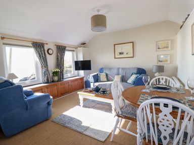 A living room with blue sofas a wooden dining table with white chairs and a mounted television at Suntrap House in Portloe