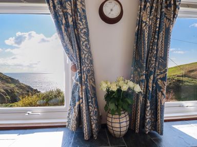 A corner with a vase of white flowers on the floor between two patterned curtains with a wall-mounted clock above with a view of hills and the sea through windows at Suntrap House in Portloe