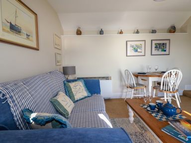 A living room with a blue checkered sofa, a wooden coffee table with a blue teapot and cups, a round dining table with four white chairs, and framed pictures on the walls at Suntrap House in Portloe