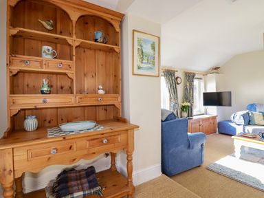 A wooden cabinet with shelves and drawers next to a living room with blue armchairs and a TV at Suntrap House in Portloe
