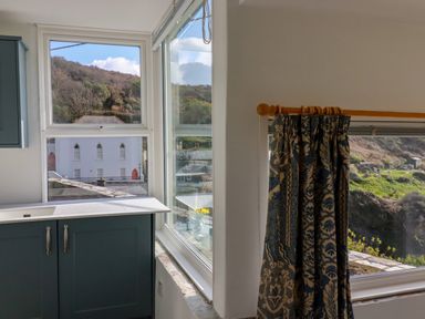 A kitchen corner with a sink under a window and a patterned curtain on a rod at Suntrap House in Portloe