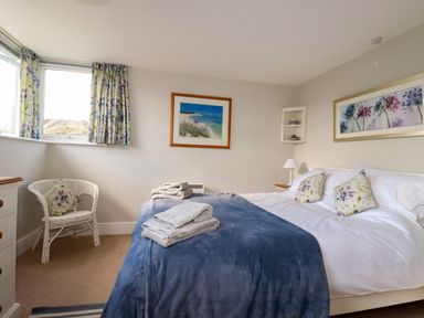 A bedroom with a bed covered with white sheets and blue blanket and towels on top a wicker chair next to a window with floral curtains at Suntrap House in Portloe