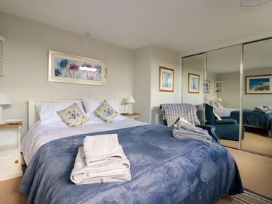 A bedroom with a double bed with blue blanket and folded towels with bedside tables and lamps and mirrored wardrobes at Suntrap House in Portloe