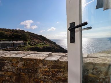 A view of a stone wall hillside houses and the sea seen through a window with a black handle at Suntrap House in Portloe