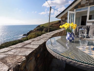 A glass table with a checkered vase of yellow flowers and a coffee press on a stone balcony overlooking the sea at Suntrap House in Portloe