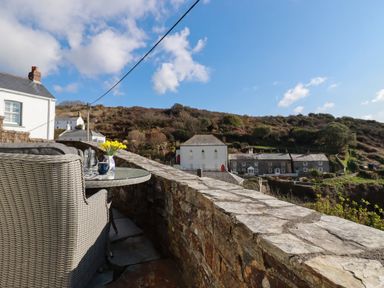 A stone balcony with wicker chairs and a glass table with flowers and tea set overlooking hills and stone houses at Suntrap House in Portloe