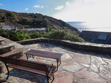 A stone patio with a wooden bench and table overlooking hillside houses and the sea at Suntrap House in Portloe