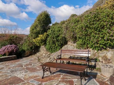 An outdoor stone patio with a wooden bench and table surrounded by bushes at Suntrap House in Portloe