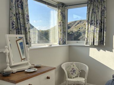 A corner of a room with a white dresser with mirror and dishes next to a white wicker chair with floral cushion at Suntrap House in Portloe