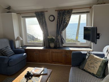A living room with two windows showing a sea view blue armchair blue sofa with cushions wooden coffee table and cabinet at Suntrap House in Portloe