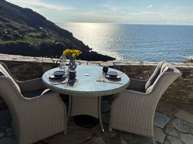 An outdoor patio with a round glass table and two wicker chairs overlooking the sea at Suntrap House in Portloe