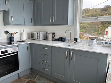 A kitchen with grey cabinets a microwave a kettle a sink and a window showing outdoors at Suntrap House in Portloe