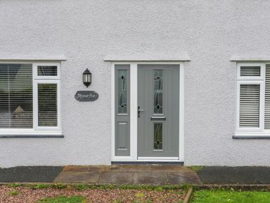 A front entrance with a gray door and two windows on a white textured wall at Monarfon Criccieth