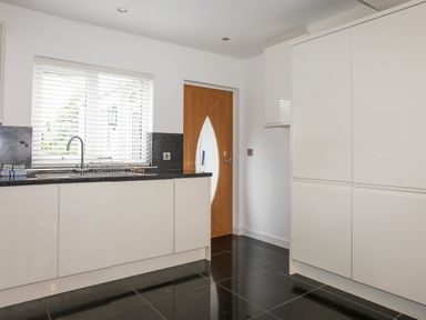 A kitchen with white cabinets black tiled floor a window with blinds and a wooden door at Monarfon in Criccieth