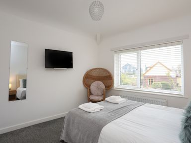 A bedroom with a bed, gray blanket, two folded towels, a wicker chair, a wall-mounted TV, a tall mirror, and a window with blinds at Monarfon in Criccieth