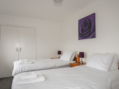 A bedroom with two single beds white bedding wooden bedside tables purple lamps and purple flower artwork on wall at Monarfon in Criccieth