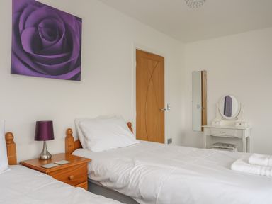 A bedroom with two single beds a wooden bedside table with a purple lamp a wooden door a white dressing table with a mirror and stool and a purple rose artwork on the wall at Monarfon in Criccieth