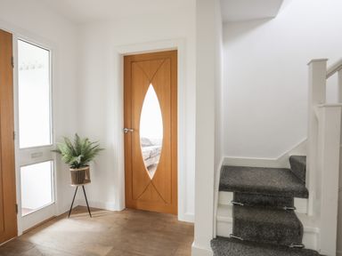 An entryway with a wooden door with an oval glass panel a potted plant on a stand a staircase with gray carpeting and a door with glass panels at Monarfon in Criccieth