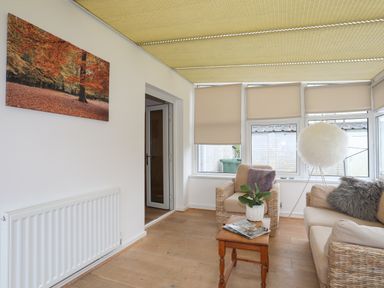 A sitting area with a small wooden table magazines a potted plant a beige armchair and sofa with cushions and lampshade in a room with windows and blinds at Monarfon in Criccieth