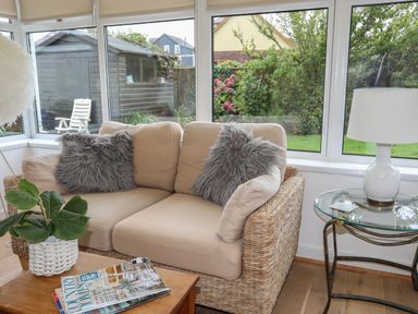 A sunroom with a wicker sofa with cushions and pillows a glass side table with a lamp a wooden coffee table with magazines and a plant at Monarfon in Criccieth