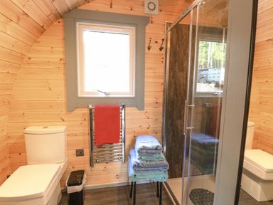 A bathroom with a toilet, window, towel rail with a red towel, stack of towels on a stool, and a glass shower enclosure at Elleric Pods - The Last Resort Fasnacloich near Appin