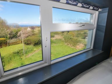 A view through a window showing a garden with grass bushes trees and a distant view of the sea at Min-Y-Ffordd in Dinas Cross