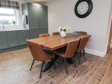 A dining area with a wooden table and six brown chairs next to green kitchen cabinets and a large wall clock at Breamish Christon Bank near Embleton