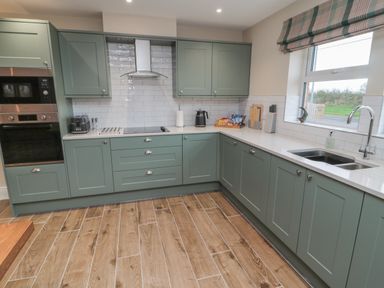A kitchen with green cabinets white countertops wooden floor an oven and microwave toaster kettle and a window with a striped blind at Breamish in Christon Bank near Embleton
