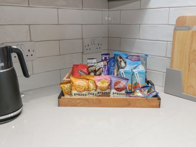 A kitchen counter with a black electric kettle a wooden tray holding various snacks and beverages and a cutting board labeled vegetables at Breamish Christon Bank near Embleton