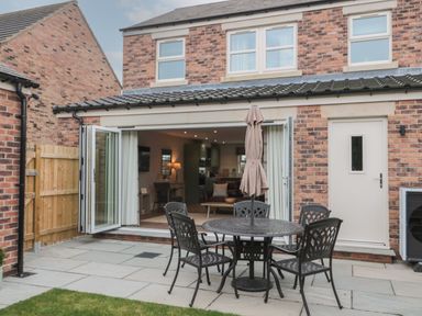 A patio with a round metal table and four chairs with an umbrella in front of a brick house with open folding doors at Breamish Christon Bank near Embleton