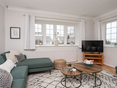 A living room with a green sectional sofa, wooden coffee tables, a television on a wooden stand, and windows with white curtains at The Palms in Abersoch