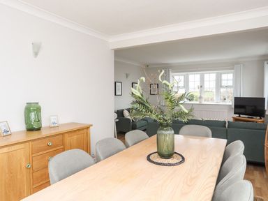 A dining room with a wooden table, grey chairs, a green vase with plants on the table, and a wooden sideboard with a green vase and photos with a living room in the background at The Palms in Abersoch