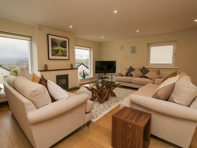A living room with three beige sofas a glass coffee table a television and large windows at Whimhowe in Windermere