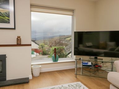 A living room with a large window showing an outdoor view and a television on a glass stand at Whimhowe in Windermere
