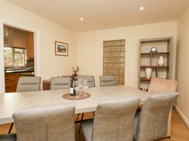 A dining room with a rectangular table surrounded by chairs a bottle of wine and glasses on the table a shelving unit with decorative items and a doorway to a kitchen at Whimhowe in Windermere