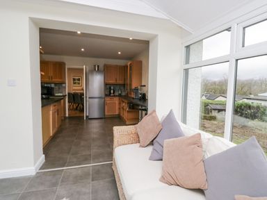 A kitchen with wooden cabinets a stainless steel refrigerator and a living area with a sofa and cushions next to large windows at Whimhowe in Windermere